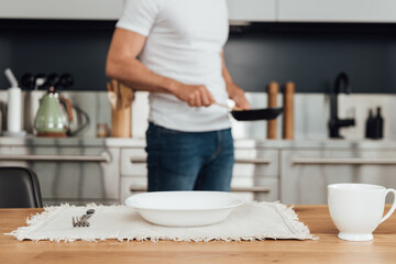 Selective focus of plate, cup and fork on table and man holding frying pan in kitchen