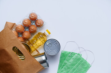 Food delivery, donation. Raw cereals, canned food, chicken eggs, sunflower oil, medical mask in paper bag on white background. Copy space, flat lay. Crisis food stock for coronavirus quarantine