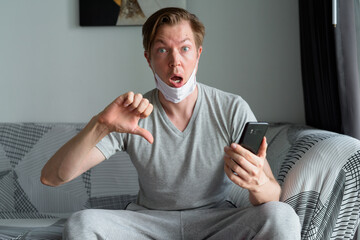 Stressed young man with mask using phone and giving thumbs down at home under quarantine
