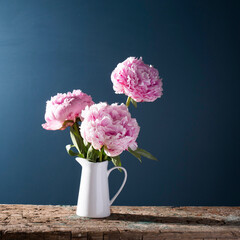 Three pink peonies in a tin jug on a shabby bench against the background of a dark blue wall