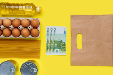 Set of products on yellow background with paper bag and euro banknotes, flatlay, top view-sunflower oil, chicken eggs, tray, canned, pasta, vermicelli, top view, food, plastic bottle, money, cash