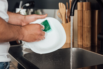 cropped view of man holding sponge while washing white plate in kitchen