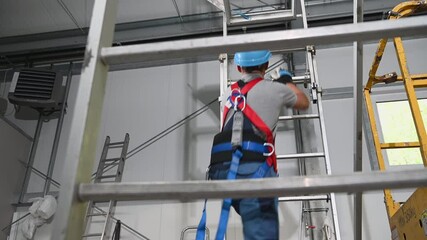 Caucasian Male Contractor Climbing Up Tall Metal Ladder Wearing Protective Harness And Hard Hat.
