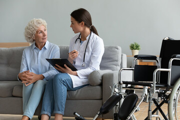 Doctor in white coat consults disabled patient explain diagnosis during homecare visit, nurse hold clipboard listen old woman fill medical form writes complaints, health care medical insurance concept