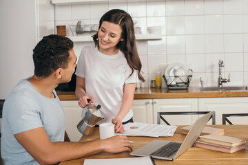 cheerful girl holding coffee pot and cup near mixed race man and laptop on table