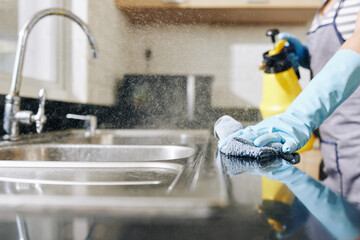 Close-up image of woman spraying disinfecting detergent on kitchen counter and sink to kill all...