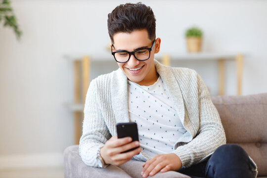 Cheerful Ethnic Male Using Smartphone On Couch.