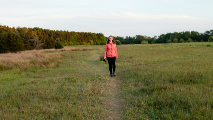 Naklejka na ściany i meble Young woman walking through a meadow carrying a camera, in late evening light.