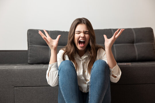 young brunette woman looks agressive hysterically shouting loud while sitting near sofa in her appartment, emotional psychology problems concept