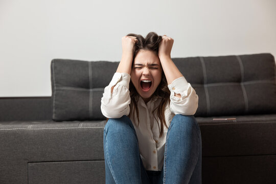young brunette woman looks exhausted hysterically shouting loud while sitting near sofa in her appartment, emotional nervous breakdown concept
