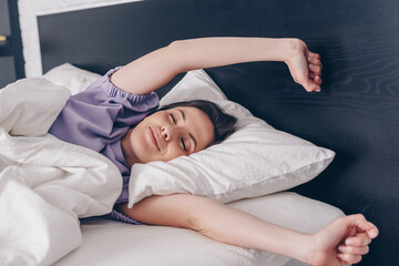cheerful young woman with closed eyes stretching in bed