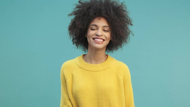 A smiling young african american woman with curly hair is posing to the camera standing isolated over blue wall background in studio