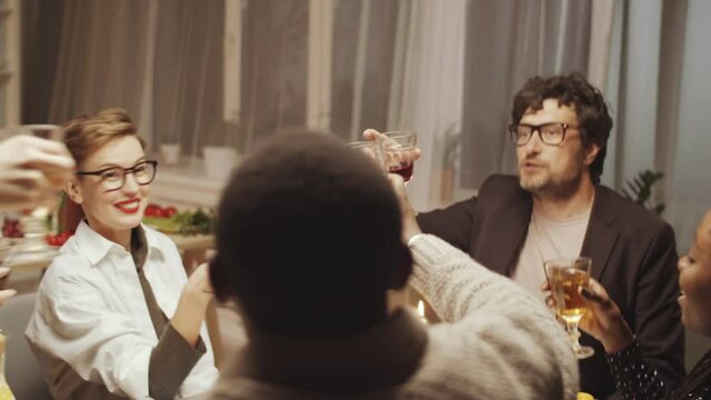Caucasian man giving a toast, clinking glasses with multiethnic friends and drinking wine at home dinner party