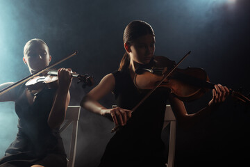 female musicians playing classical music on violins on dark stage with smoke