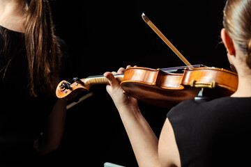back cropped view of two female professional musicians playing on violins on dark stage