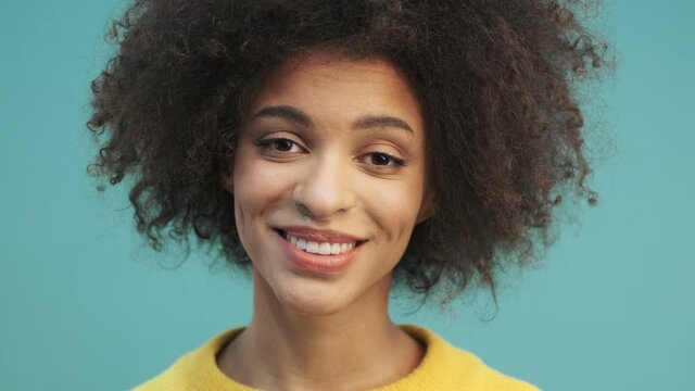 A close-up view of a beautiful young african american woman with curly hair is posing standing isolated over blue wall background in studio