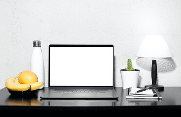 Workplace of freelancer, laptop with mockup on black desk beside lamp, steel thermo water bottle, pen on notebook, fruits and green cactus pot. Background of grey textured wall.