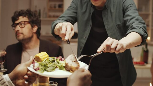 Young hospitable man using knife and fork while putting baked meat on plates for his friends at home dinner party