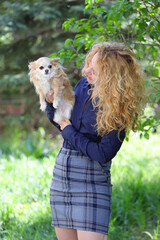 Blond young woman, business dressed, holding a long-haired chihuahua dog outdoor, on a green natural background.
