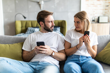 Young couple using smartphones in living room at home. Jealous boyfriend spying his girlfriend's phone.