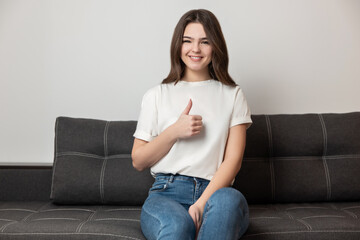 young beautiful brunette woman sitting on the sofa in her appartment showing like sign looking...