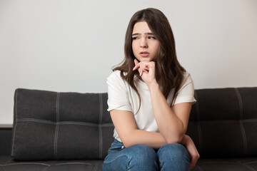 Obraz premium young beautiful brunette woman sitting on the sofa in her appartment holding hand near chin looking upset, feels cozy at home