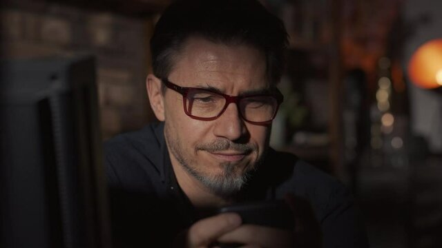 Portrait of good looking man in glasses at home in a dark room using smart phone and thinking, looking at the camera and smiling at the end. 4K video footage.
