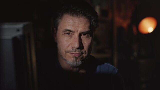 Portrait of a handsome man in a dark room leaning forward into the light while looking at the camera and leaning back to the dark at the end. High quality 4K video footage.