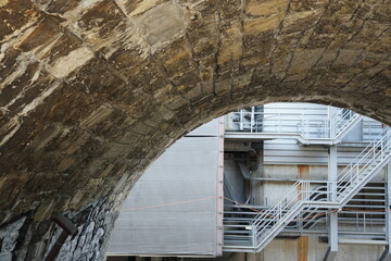 arch made of stone cubes giving view to a back tract of an industrial buildings with pipes and metal staircase