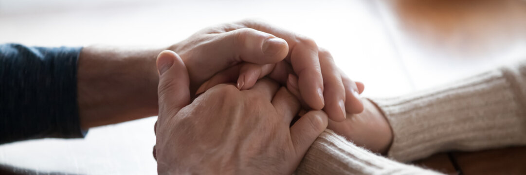 Middle-aged couple sit indoors holding hands close up photo. Spouses feeling connection and love, share problems express empathy showing compassion concept. Horizontal banner for website header design
