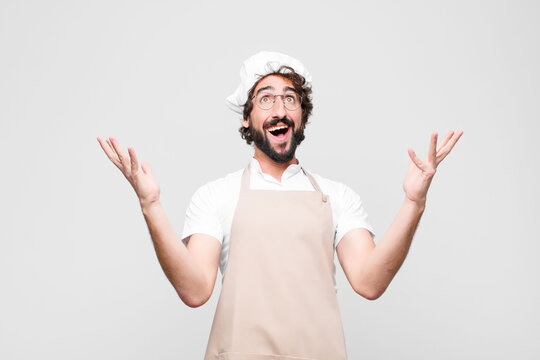 young crazy chef feeling happy, amazed, lucky and surprised, celebrating victory with both hands up in the air against white wall