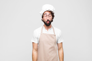 young crazy chef pressing lips together with a cute, fun, happy, lovely expression, sending a kiss against white wall