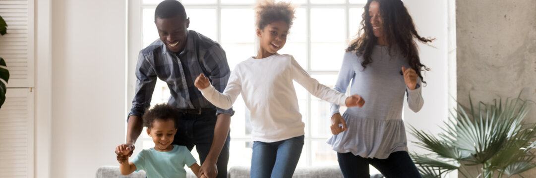 African couple and little kids dancing in living room. New first dwelling moving day celebration, active weekends with cute children at home concept. Horizontal photo banner for website header design