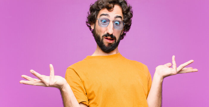 young crazy cool man shrugging with a dumb, crazy, confused, puzzled expression, feeling annoyed and clueless against flat wall