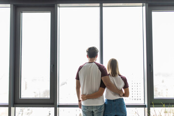 Young beautiful couple stand near window embrace. Happy smile man and woman lovers hug. Back rear view