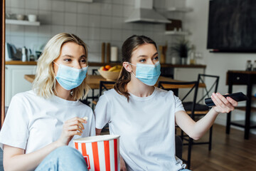 Fototapeta na wymiar Sisters in medical masks holding popcorn while watching tv at home
