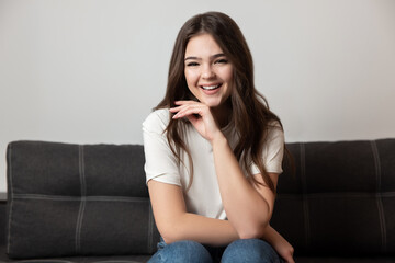 portrait of young beautiful smiling brunette woman sitting on the sofa in her appartment holding hand near face looking happy, feels cozy at home