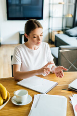 attractive woman holding bottle with hand sanitizer near notebook and cup on table