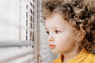 Portrait of a pretty curly girl in a bright yellow dress standing near a light window with blinds