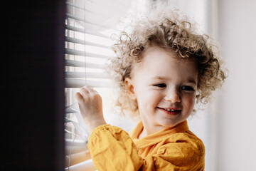 Portrait of a pretty curly girl in a bright yellow dress standing near a light window with blinds