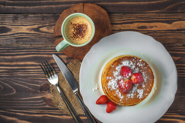 Pancakes with strawberries, icing sugar and a cup of cappuccino for breakfast on a wooden background in a rustic style. top view