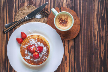 Pancakes with strawberries, icing sugar and a cup of cappuccino for breakfast on a wooden background in a rustic style. top view