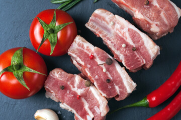 Raw slices of pork on a black slate board, tomatoes, red pepper and powder. Pork belly with vegetables.