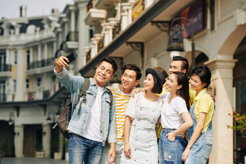 Obraz premium Happy young Vietnamese man taking selfie with friends when they are walking in historical center of city