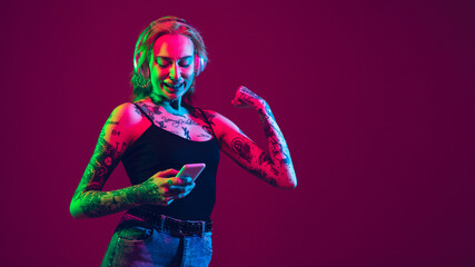 Listening to music with wireless headphones, scrolling phone. Portrait of young caucasian woman on purple background in neon. Human emotions, facial expression, sales, ad concept. Flyer, copyspace.