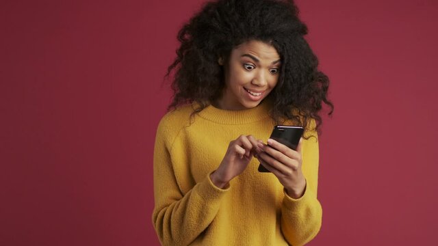 Young african shocked woman isolated over dark red burgundy background using mobile phone