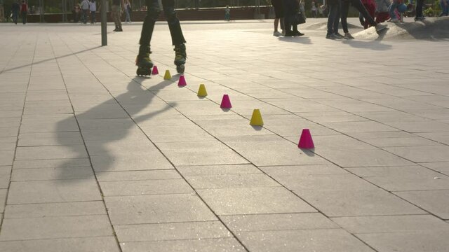 Close-up legs of a young man is professionally skating between cones on a nice evening sunset in a city park.