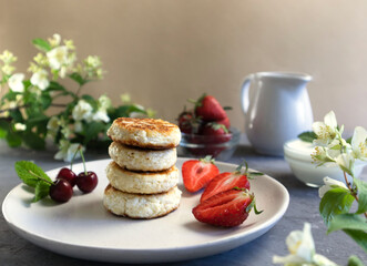 cheese pancakes or curd pancakes with strawberries and cherries, the concept of a healthy breakfast, in the background a jug of cream and bouquets of jasmine, horizontal orientation