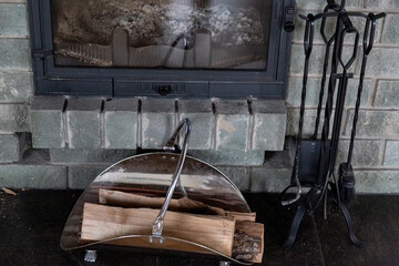 pine firewood on an iron stand and cast-iron accessories like a poker and a scoop on a background of a gray brick fireplace