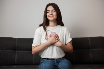 brunette woman looks satisfied holding her smartphone in oher hands with love and care sitting on the sofa in her appartment , technology era concept